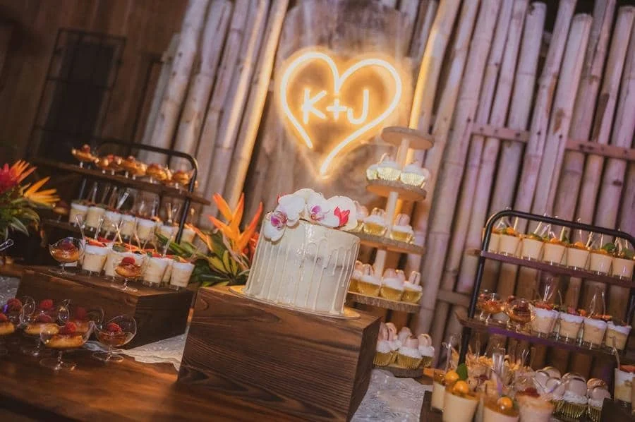 wedding reception and party photography in costa rica -8 | Pacific Photography Dessert table at a rustic wedding: a white frosted cake with orchids on a wooden pedestal, surrounded by cupcakes and pastries, with a neon heart sign reading K+J in the background